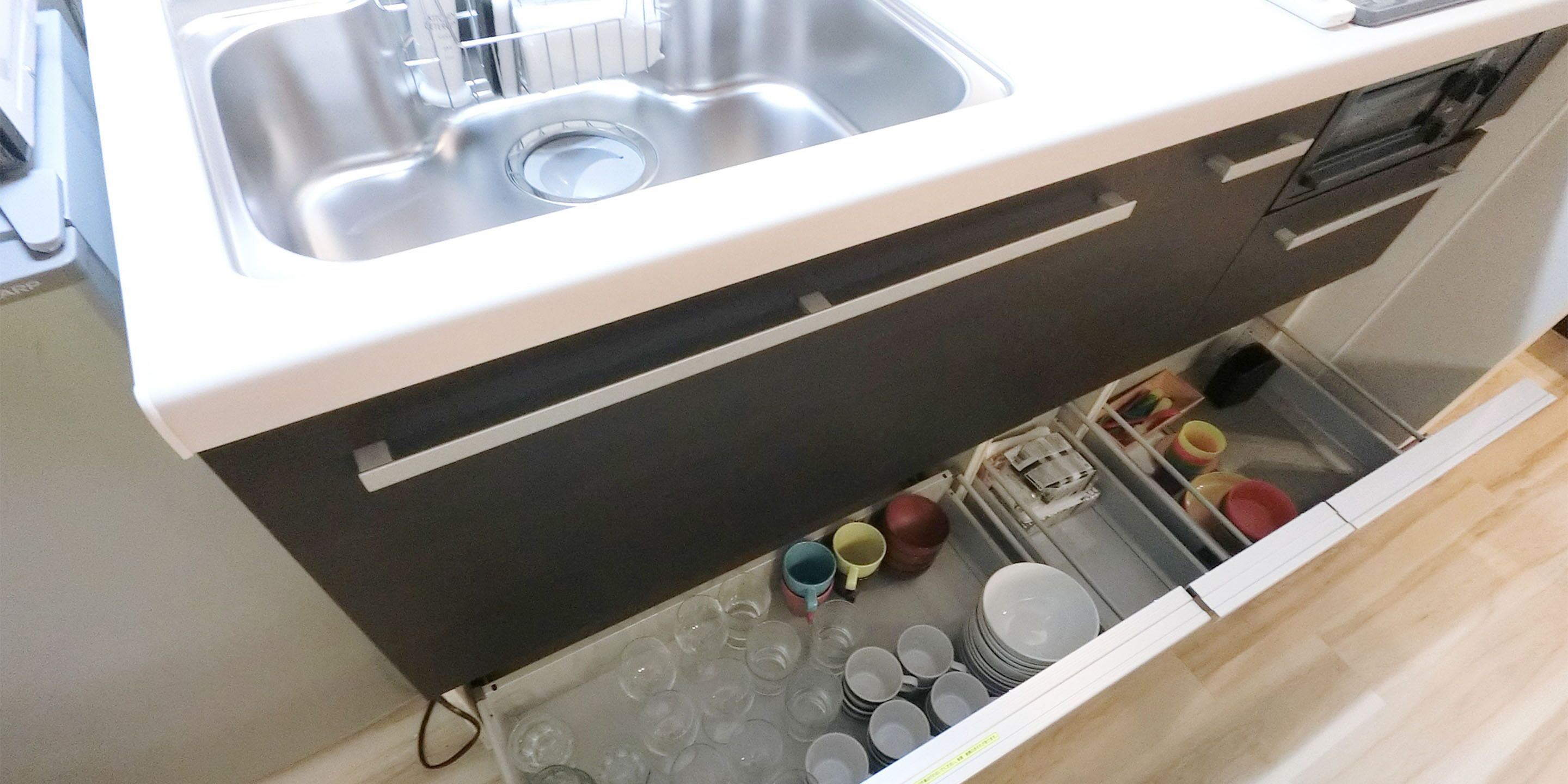 A kitchen with a stainless steel sink, a white countertop, and a drawer below containing various dishes and glasses.