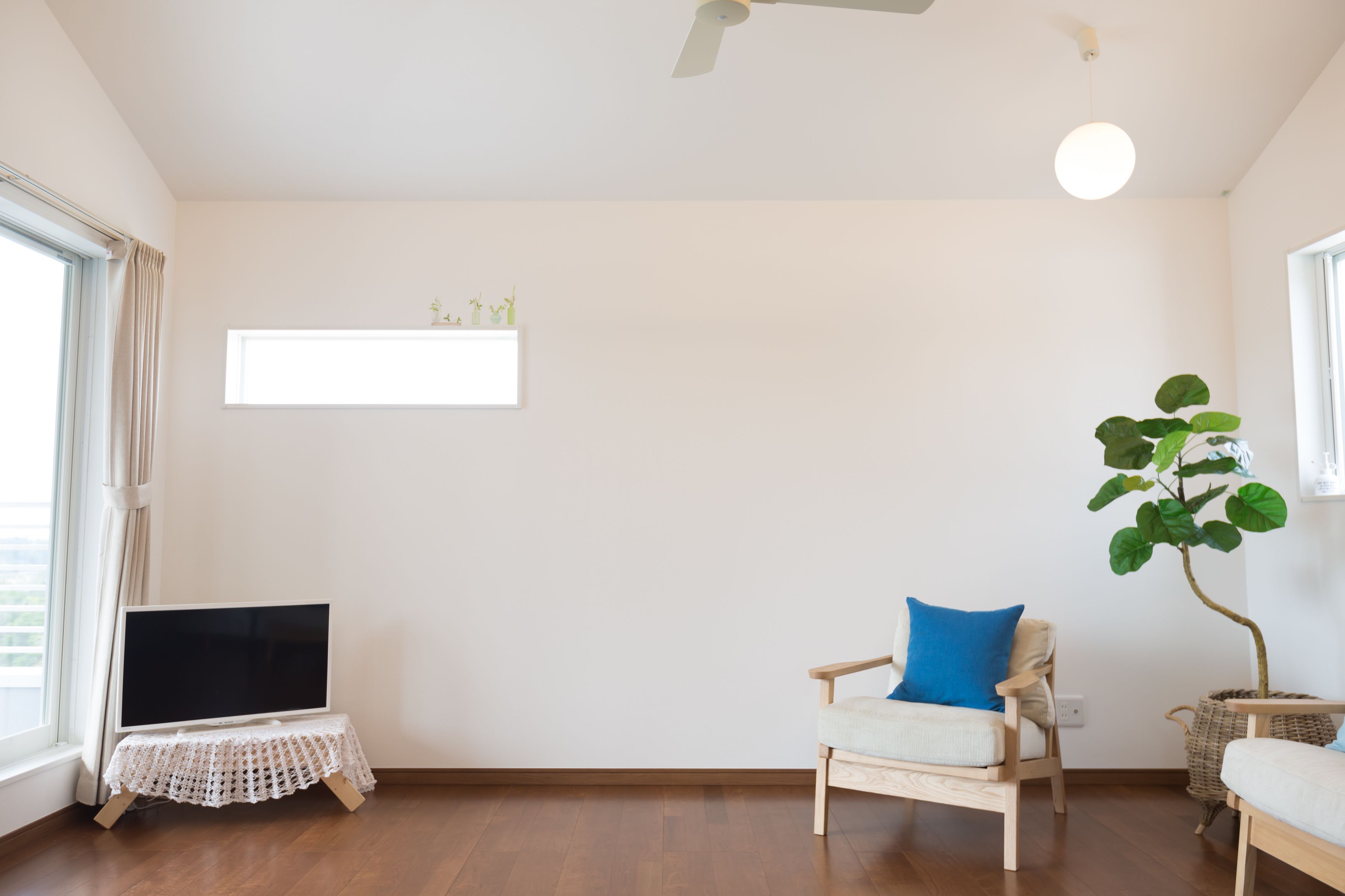 A living room with a flat-screen TV on a small table, a chair with a blue pillow, a large plant, and a ceiling fan.