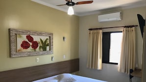 Una habitación de hotel con una cama, un cuadro de rosas rojas, un equipo de aire acondicionado y una ventana con cortinas.