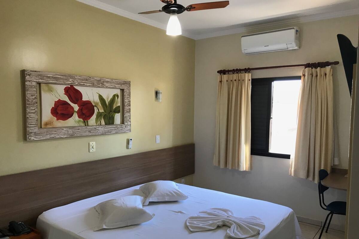 A hotel room with a bed, a painting of red roses, an air conditioning unit, and a window with curtains.