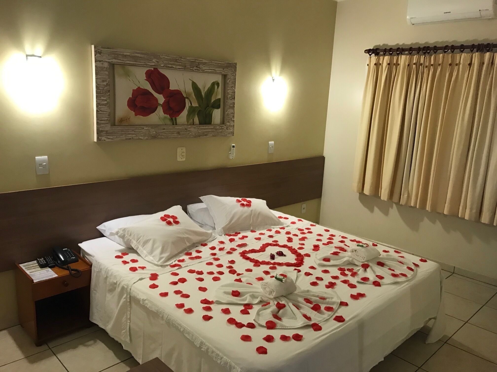 A hotel room with a bed adorned with rose petals and a heart-shaped arrangement, a painting of red roses on the wall, and a telephone on the nightstand.