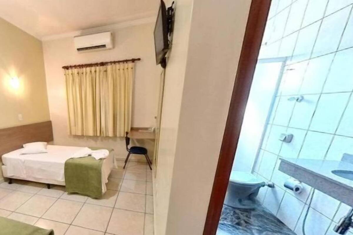 A hotel room with two single beds, a small table, a chair, an air conditioning unit, and a view of the outside balcony.