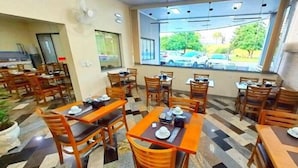 Un restaurante con mesas y sillas de madera, piso de baldosas y vista a coches estacionados a través de amplios ventanales.