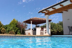 A swimming pool with a tiled edge, a wooden pergola, and a white building with a tiled roof.