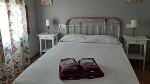 En pænt redt seng med et hvidt sengegærde, to sengeborde med lamper og en blomstret gardin.