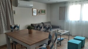 Et spiseområde med et træbord og stole, en lille plante på bordet, en sofa og et sofabord. Der er to blå fodskamler og et vindue med gardiner.