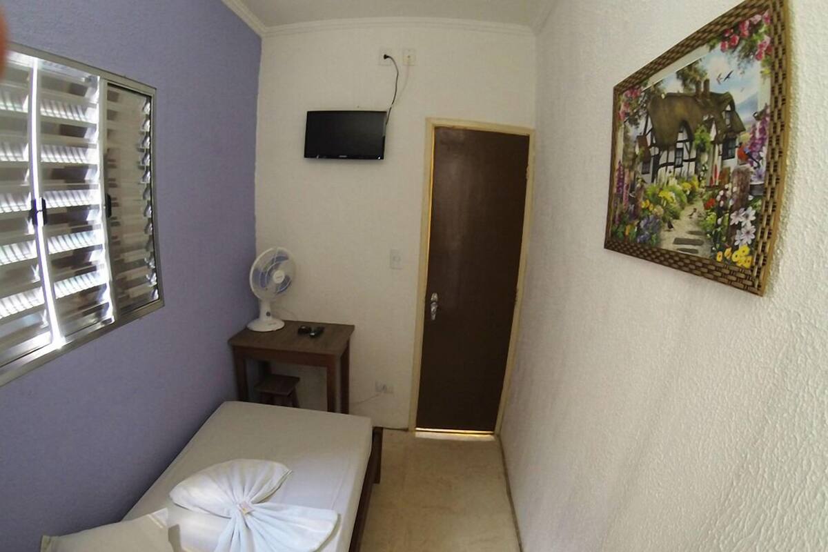 A narrow hallway with a bed on the left, a small table with a fan, a TV mounted on the wall, and a door at the end.