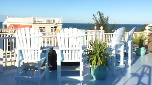 A terrace with white Adirondack chairs and potted plants, overlooking a coastal view.