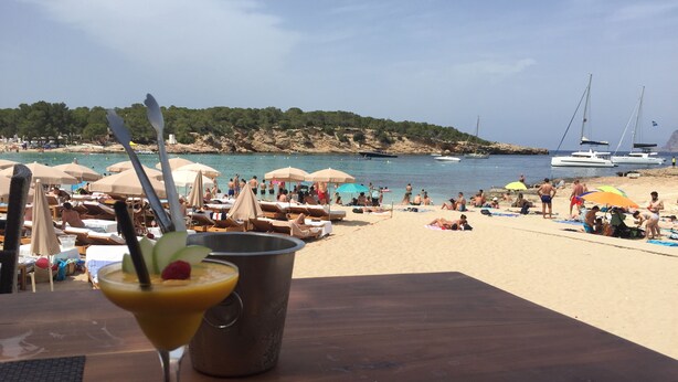 A beach scene with umbrellas, lounge chairs, and a table with a drink.