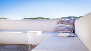 A rooftop terrace with a white cushioned bench, a colorful woven pillow, and a white table.