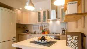 A modern kitchen with light wood cabinets, a central island with a bowl of fruits and vegetables, and a decorative panel on the right. - La Vue Sur La Mer Beach House (Viareggio)