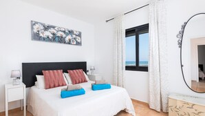 Um quarto com cama, mesas de cabeceira, um quadro, janela com cortinas e vista para o mar.