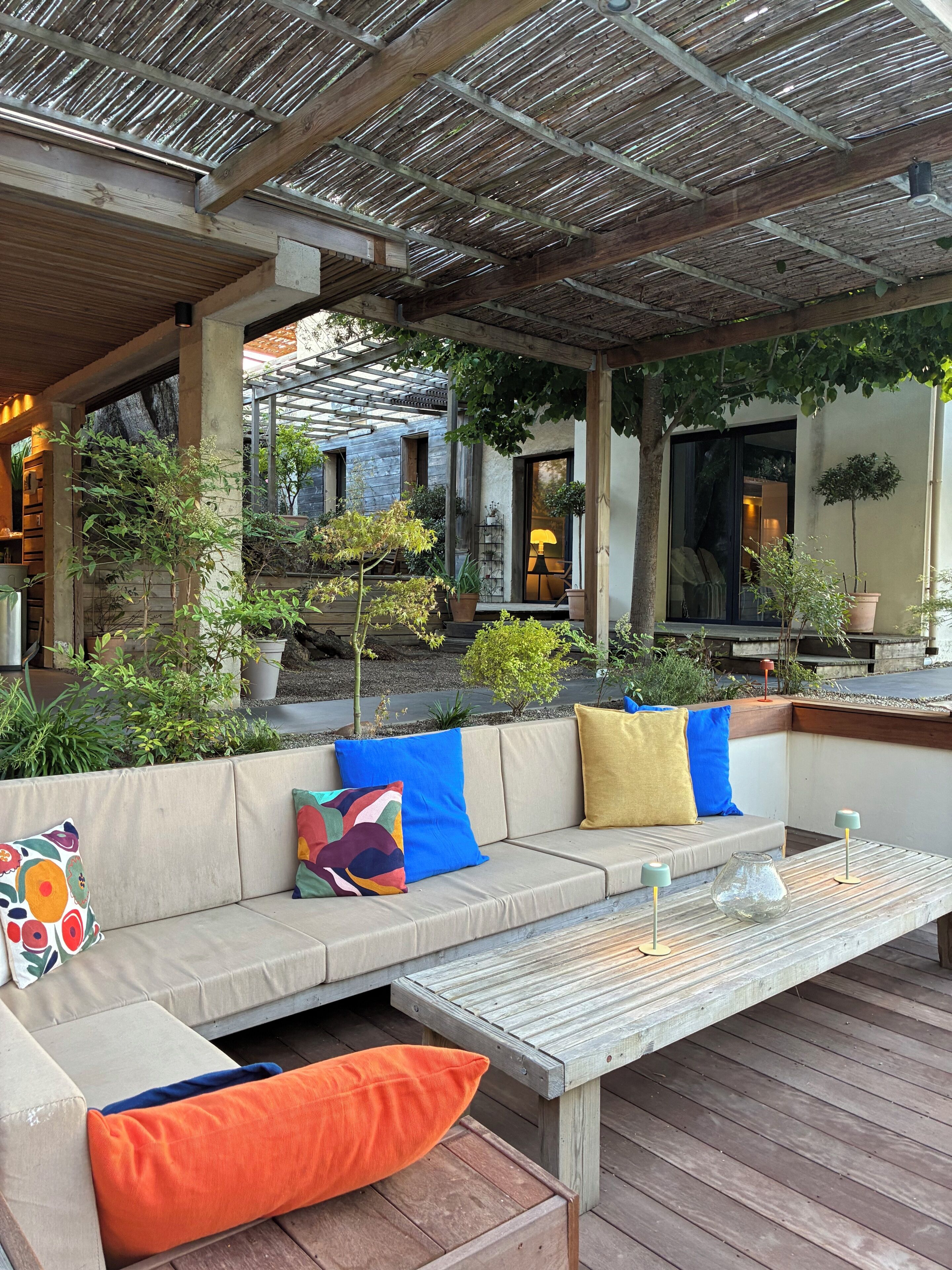 Un espace extérieur aménagé avec un banc en bois et des sièges rembourrés, entouré de plantes en pot et d’une structure en forme de pergola.