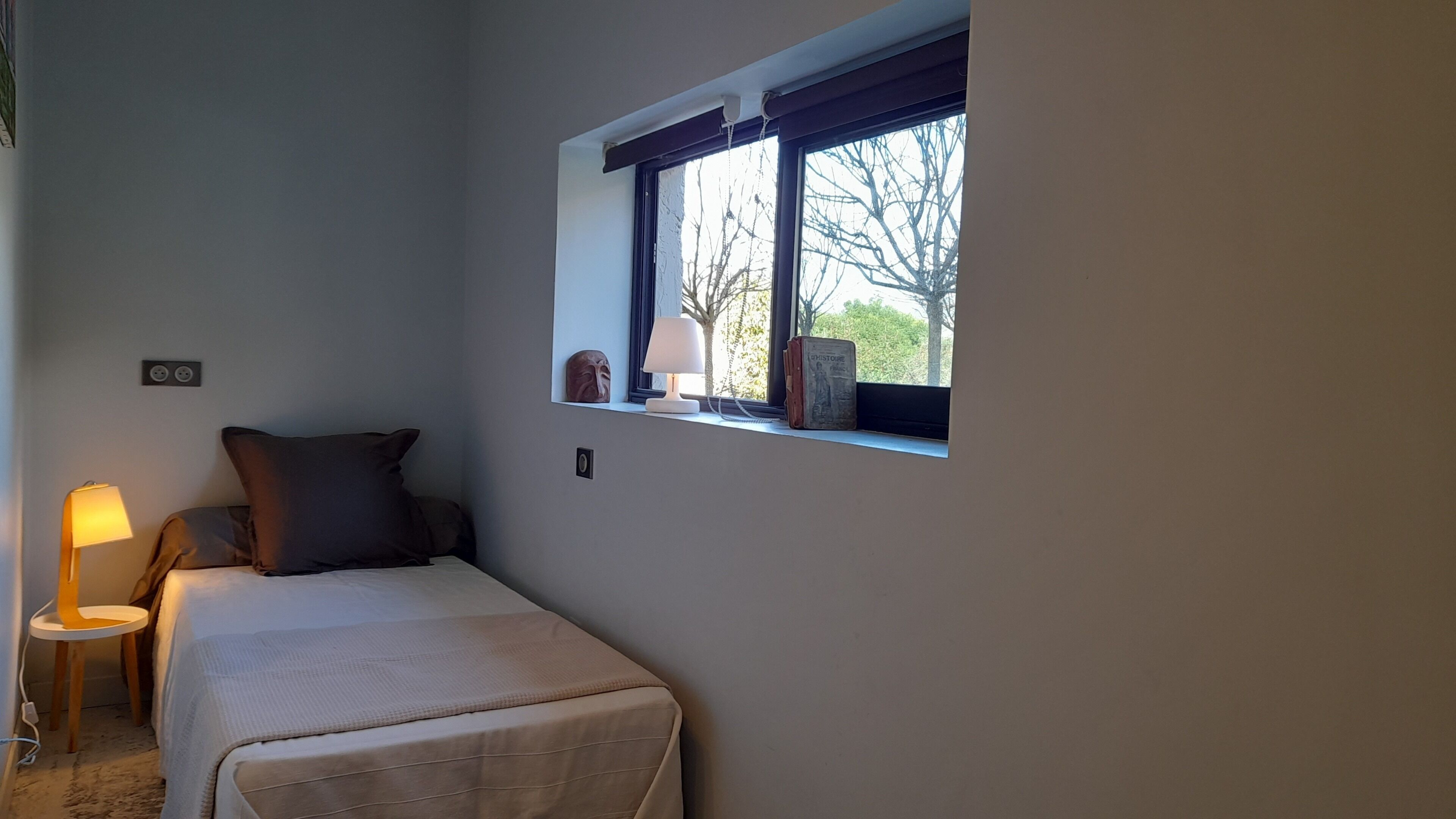 A small, neatly arranged bedroom with a single bed, a bedside lamp, a window with a view of trees, and a small table with a lamp.