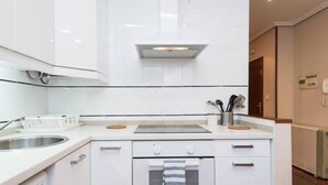 Una cocina moderna con armarios blancos, un lavamanos de acero inoxidable y una campana extractora integrada. Se observa una pequeña ventana sobre el lavamanos y un refrigerador blanco a la derecha.