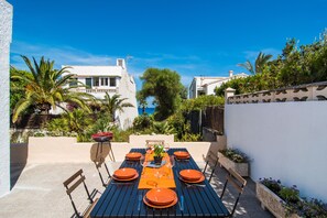 Un espace de restauration en plein air avec une table dressée pour six personnes, entouré de verdure et offrant une vue sur la mer.