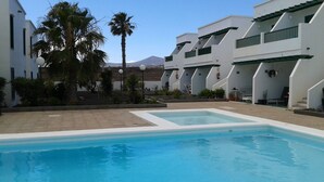 A swimming pool surrounded by white buildings with green accents, palm trees, and a mountain in the background.