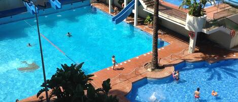 A large indoor swimming pool with multiple lanes and a slide, surrounded by palm trees and a tiled deck area.