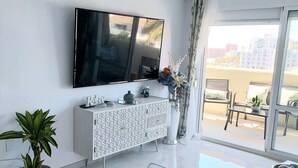 En moderne stue med et fladskærms-tv på væggen, et hvidt konsolbord med et mønster, et sofabord i glas, og udsigt til det fri gennem et stort vindue.
