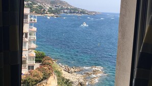 Vista costera desde una ventana con vista a un litoral rocoso, edificios y un mar azul cristalino.