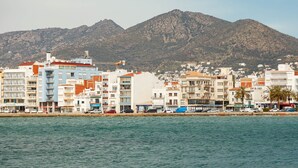 Un paisaje urbano costero con edificios de varios pisos, una montaña al fondo y un mar tranquilo en primer plano.