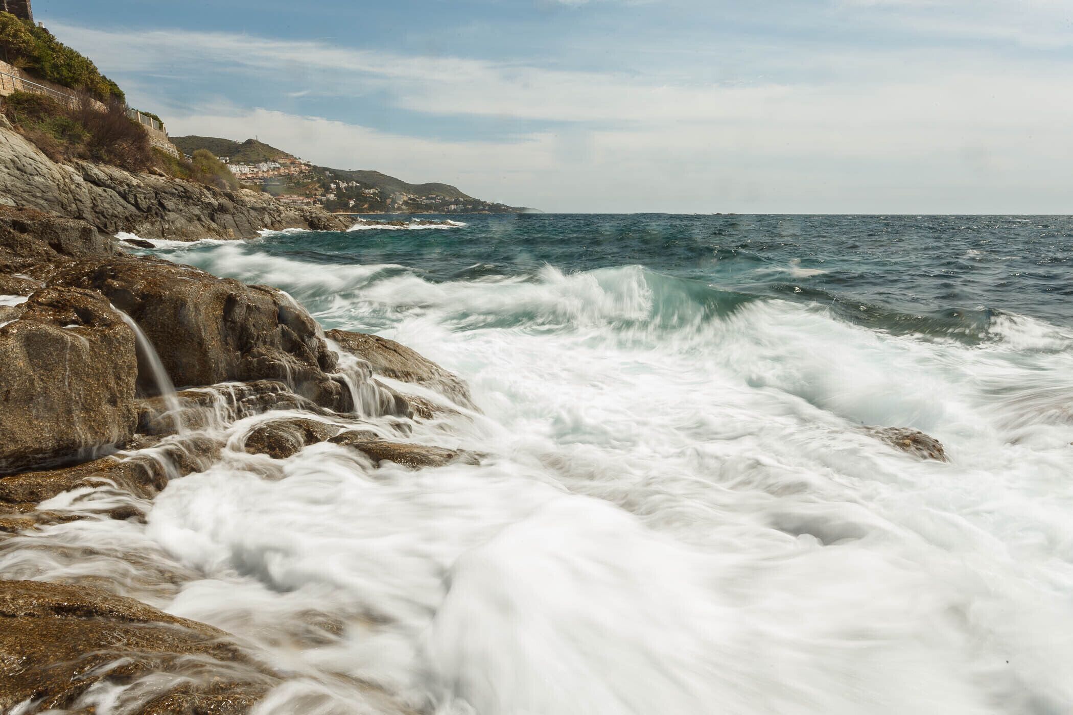 A coastal scene with waves crashing against rocks, a clear sky, and a distant view of a town on a hill.