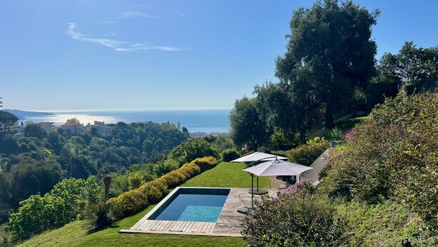 A swimming pool with a wooden deck, surrounded by lush greenery and a view of the sea.