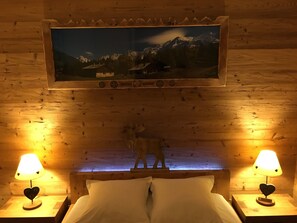 A wooden cabin room with a bed, two bedside lamps, a mounted picture of a mountain landscape, and a wooden headboard.