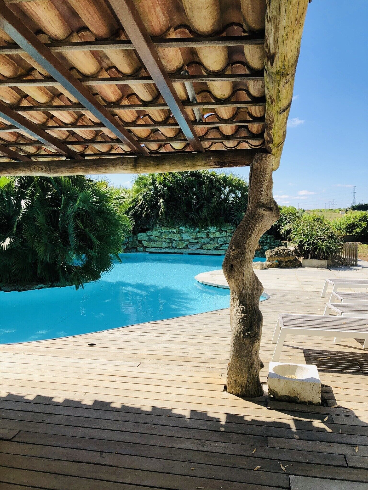 Une terrasse en bois avec une piscine, entourée de verdure et d’un mur de pierres.