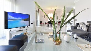 Uma sala de estar moderna com televisão de tela plana, um sofá, uma mesa de centro com objetos decorativos e uma área de jantar ao fundo.