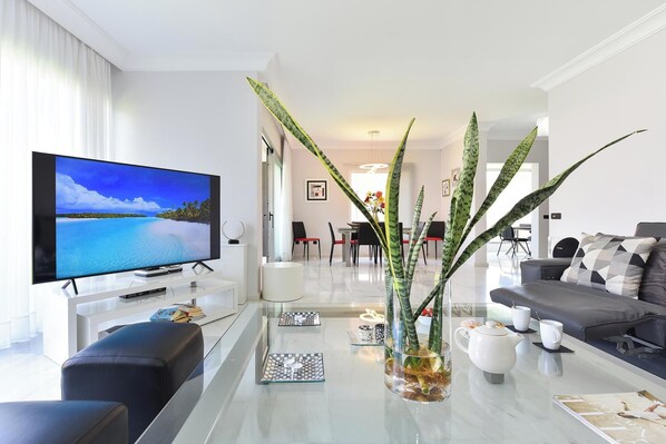 Een moderne woonkamer met een flatscreen televisie, een bank, een salontafel met decoratieve objecten, en een eethoek op de achtergrond.