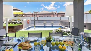Mesa de jantar posta para uma refeição, com vista para uma piscina e casas ao fundo.
