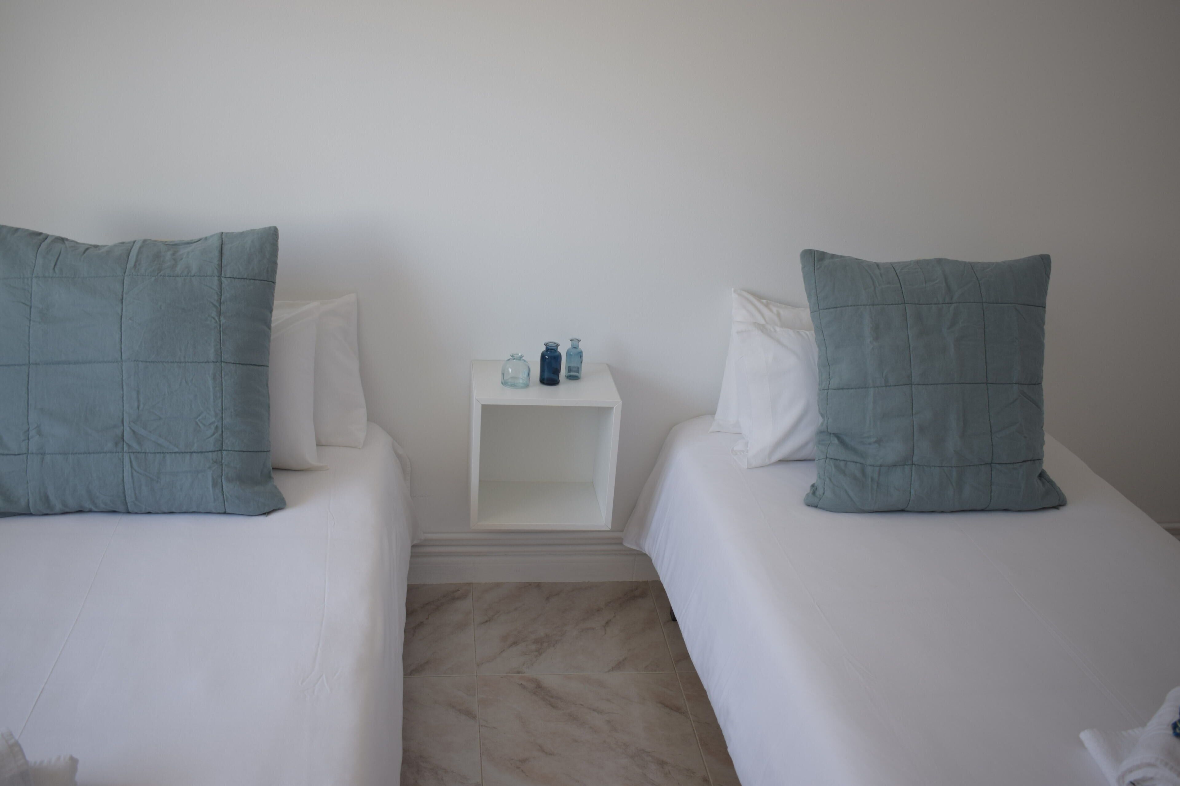 Una habitación con dos camas, cada una con un cojín verde agua y uno blanco. Hay una mesita de noche pequeña con algunos objetos encima.