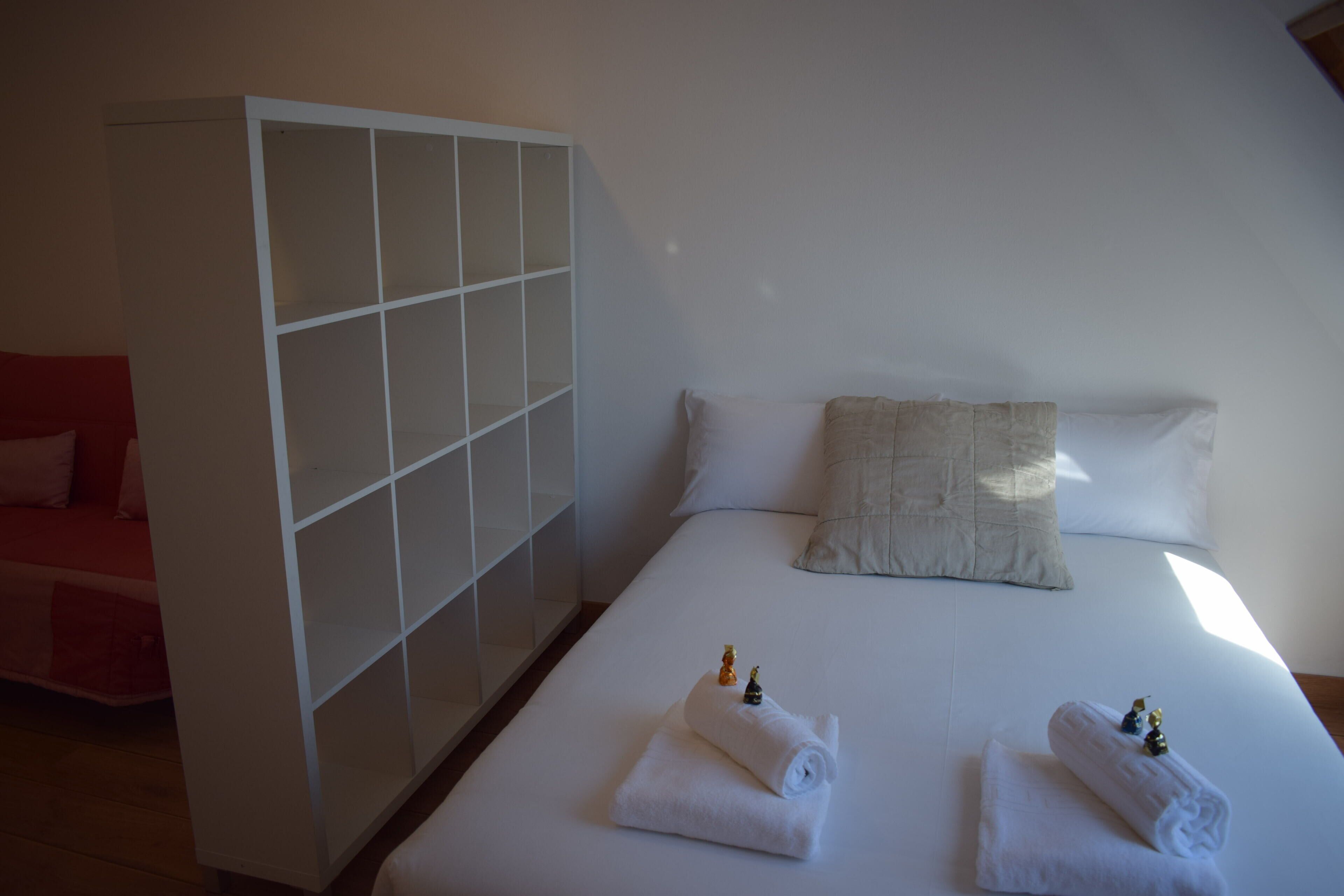 Hotellihuone, jossa on sänky, kaksi tyynyä, valkoinen vaatekaappi ja kaksi rullalle taitettua pyyhettä, joiden päällä on koriste-esineitä.