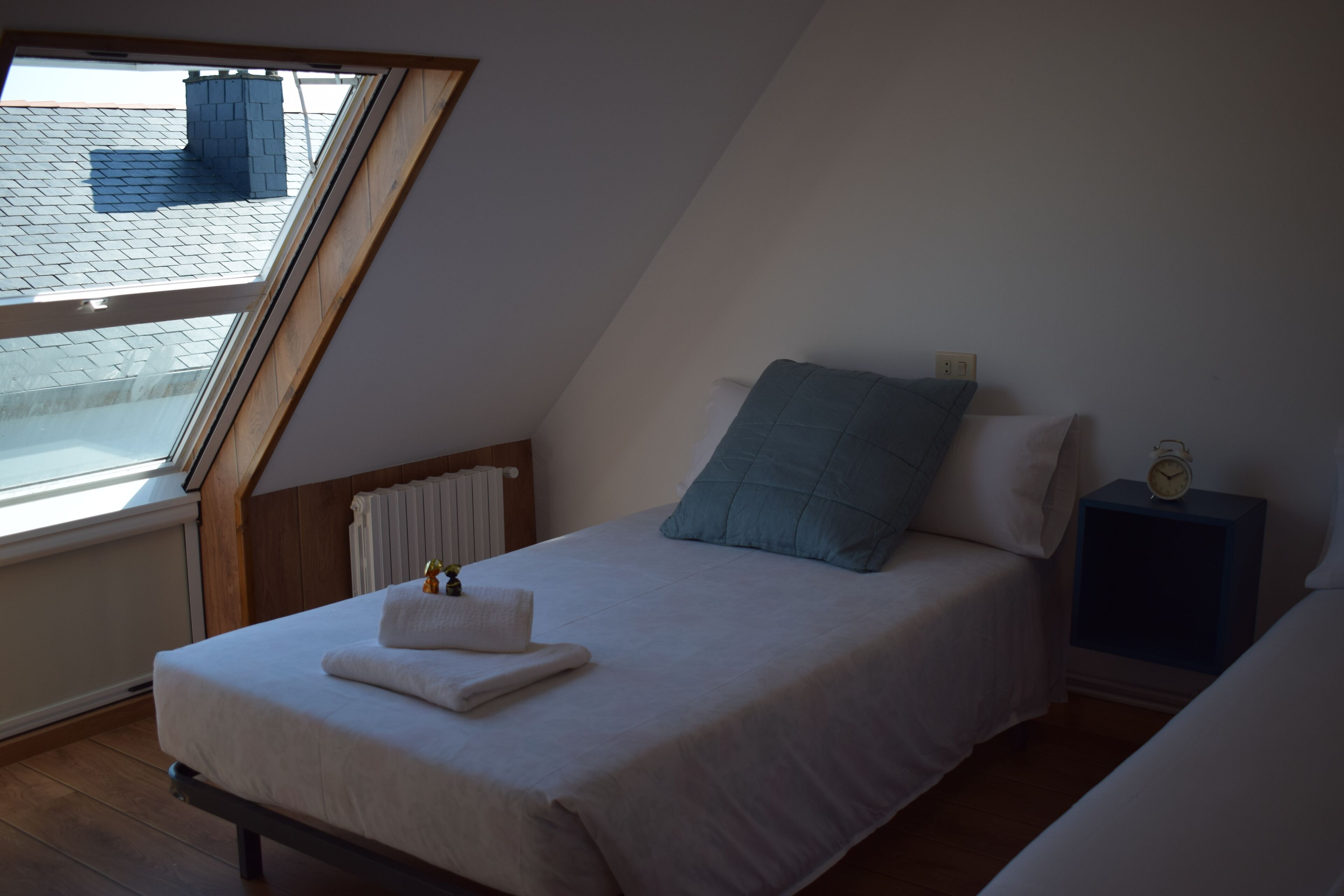 Un dormitorio con techo inclinado, una cama con ropa de cama blanca, un almohón azul, una mesita de noche con un reloj y una ventana con vistas a un tejado.