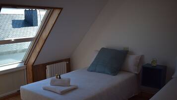 Un dormitorio con techo inclinado, una cama con ropa de cama blanca, un almohón azul, una mesita de noche con un reloj y una ventana con vistas a un tejado.