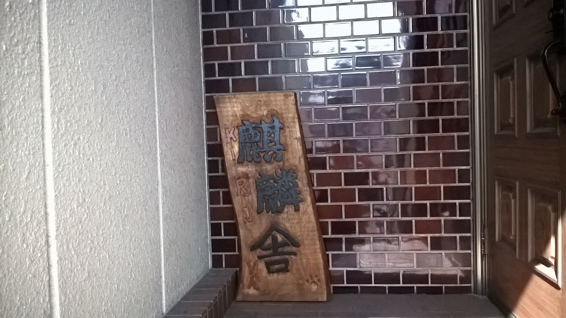 A wooden plaque with engraved text in a corner of a room with a tiled wall and a door.