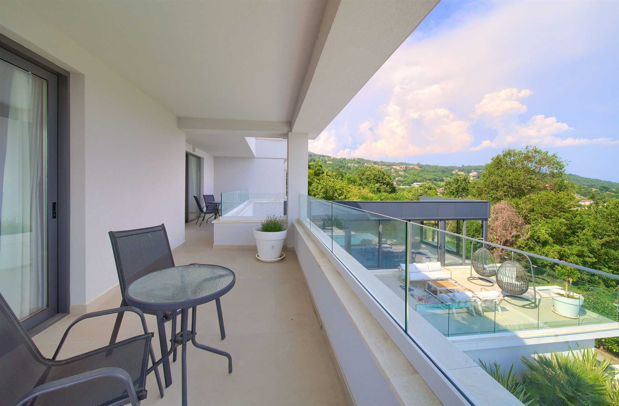 Un balcón con barandilla de vidrio, una mesa con sillas, una planta en maceta y vista a un paisaje verde.