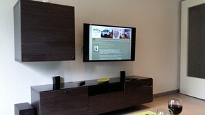 Una sala de estar moderna con un televisor de pantalla plana montado en la pared, un mueble de madera oscura debajo y una mesa de centro verde con dos copas de vino.