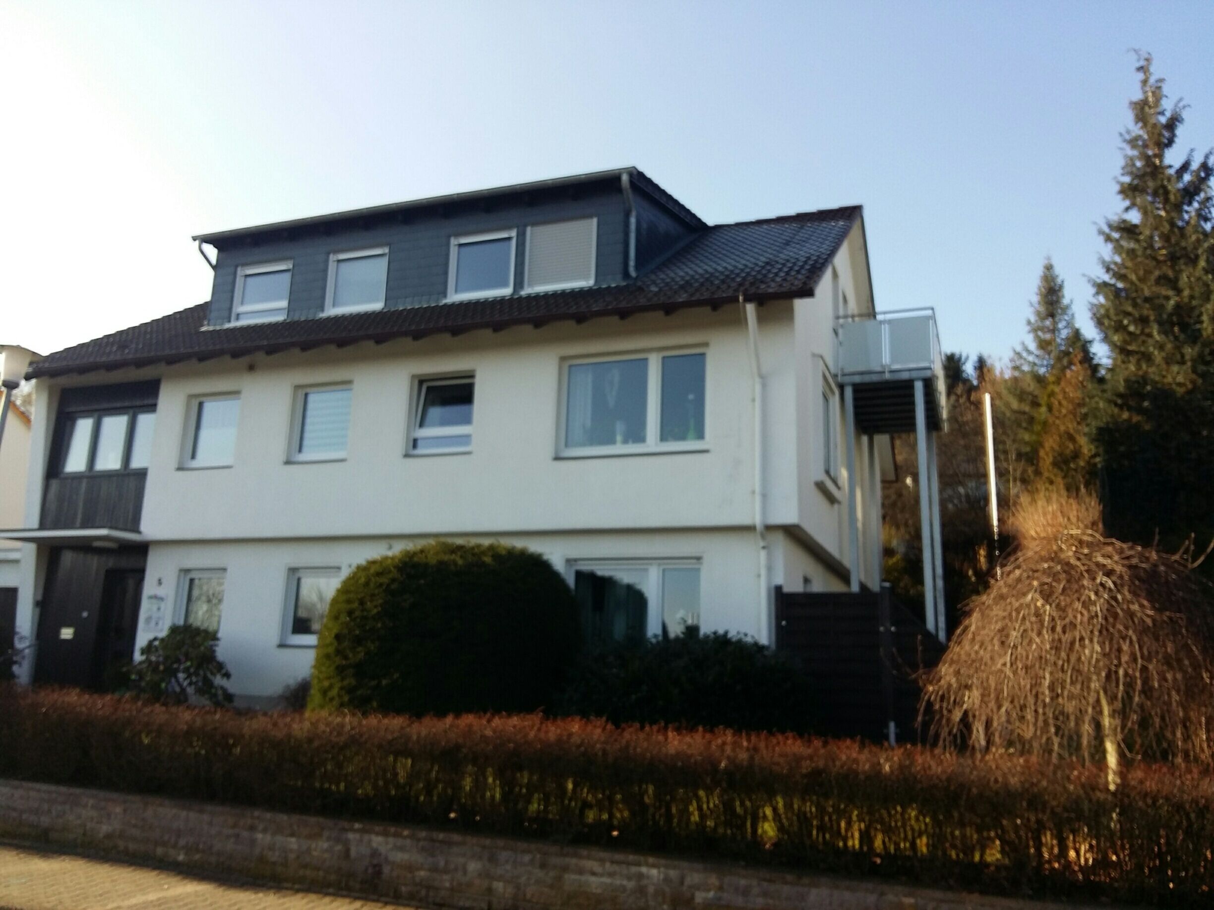 A two-story residential building with a flat roof, multiple windows, and a small garden area with a hedge and a tree.