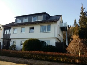 A two-story residential building with a flat roof, multiple windows, and a small garden area with a hedge and a tree. - Self-in Goslar, suitor Apartments - Attic (Goslar)