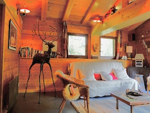 A cozy cabin interior with a wooden ceiling, a white sofa with pink pillows, a deer sculpture, and a bookshelf.