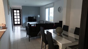 A modern dining area with a white table, black chairs, and a decorative vase. A television is mounted on a cabinet against a wall with a window.