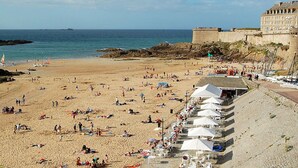 A beach with a row of white umbrellas and beach chairs along the shore, a sandy beach, and a stone wall with a building in the background.
