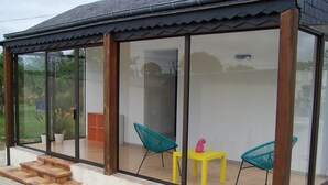 A modern glass-enclosed patio with a yellow table and two chairs, a wooden staircase, and a view of a garden.