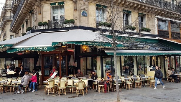 An outdoor cafe with tables and chairs, a sign reading "LES DEUX MAGOTS," and a building with ornate architecture in the background. - HISTORIC CENTER St. Germain-Près—close to the metro, the river, shopping, comfortable, quiet, and bright (Paris)