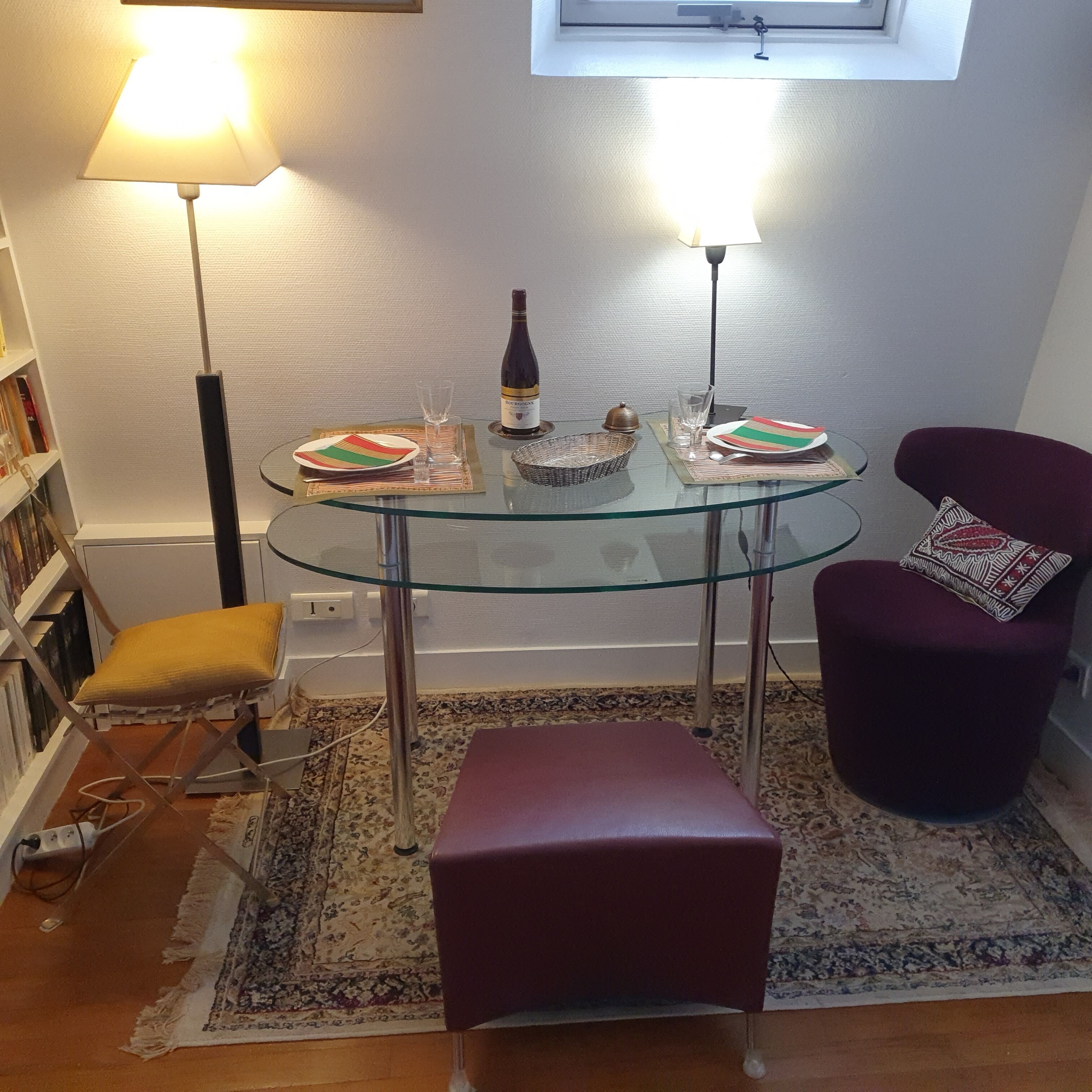 A small, modern living room with a glass-top table set for two, a purple armchair, a yellow cushion, a red ottoman, and a floor lamp.