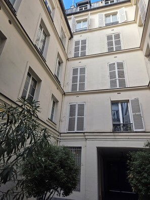 Un immeuble à plusieurs étages avec des volets blancs aux fenêtres, une porte bien visible et un arbre au premier plan.