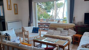 Un salon chaleureux avec un canapé en bois, une table basse et un téléviseur. Il y a une fenêtre avec des rideaux, un luminaire au plafond et une vue sur les arbres à l’extérieur.