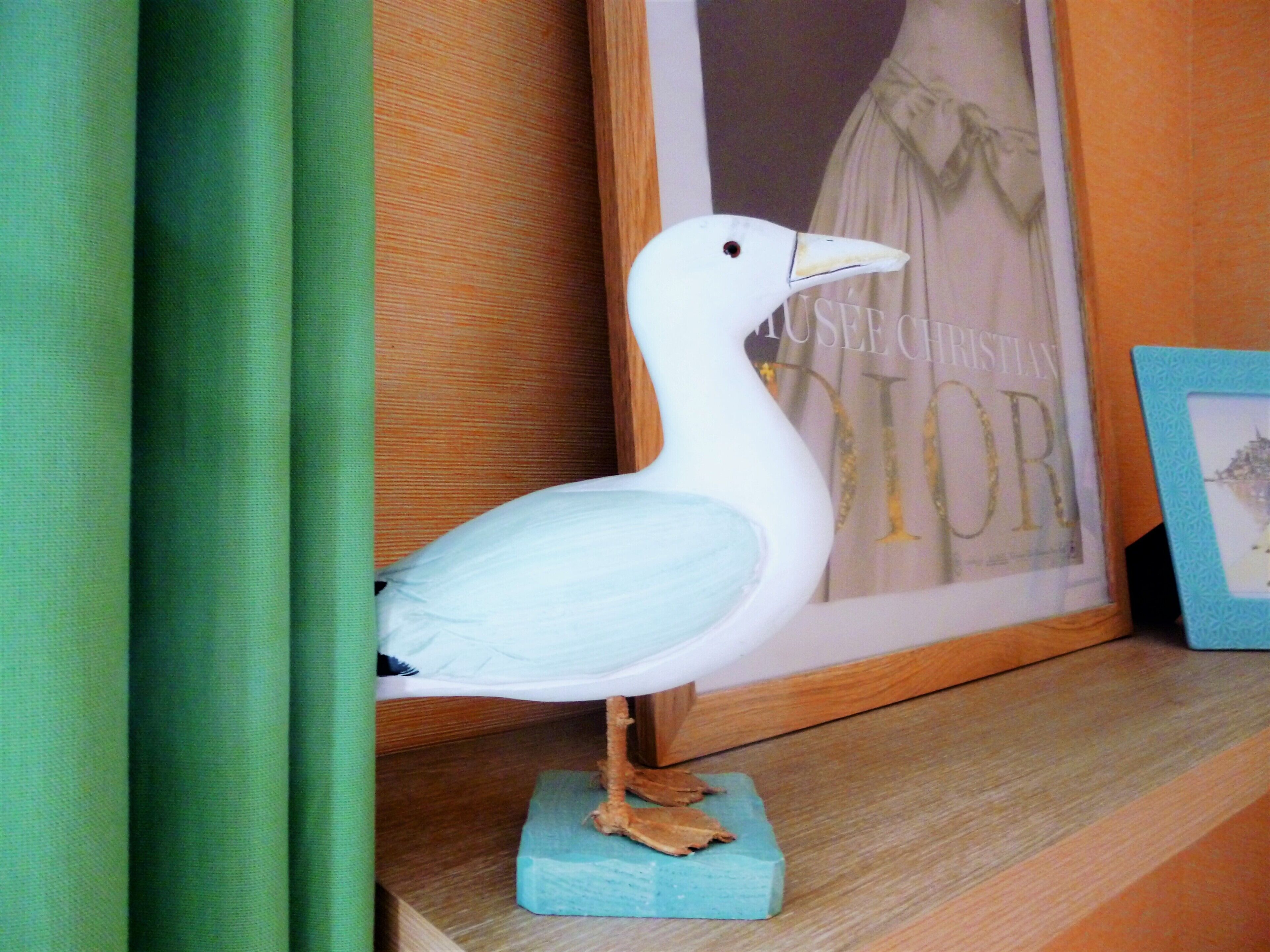 A white seagull figurine on a blue base, positioned on a wooden shelf with a framed picture in the background.
